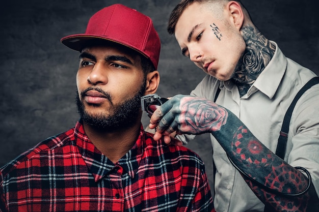 Free photo a tattooed caucasian male dressed in a white shirt cutting the beard of black hipster males in a red fleece shirt.