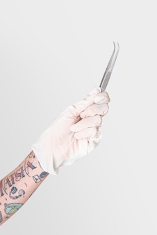 Татуированная рука в белой перчатке с изогнутым пинцетом