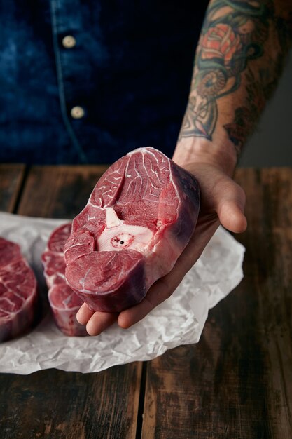 tattooed hand offers a piece of meat stake above two steaks on craft paper, close up