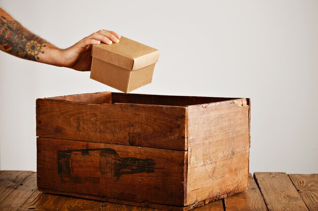 Tattooed hand picks up cardboard package from old wooden crate on rustic table, isolated on white