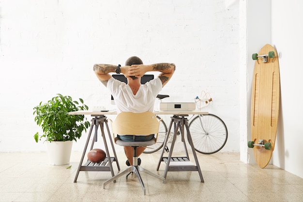 Free photo tattooed man in blank white t-shirt looks in monitor with his hands folded behind head back view in big loft room with brick wall and longboard, rugby ball, green plant and vintage bicycle around him