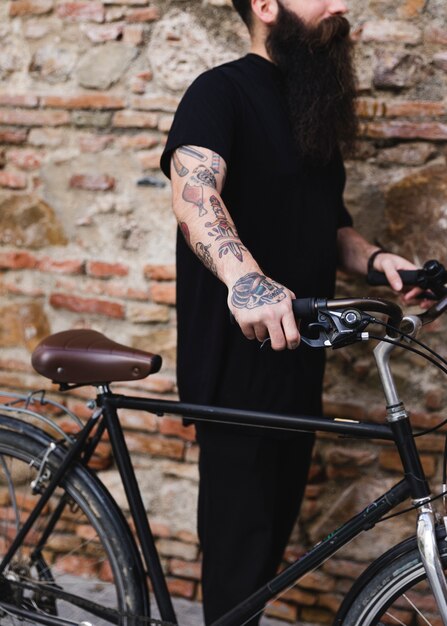 Tattooed man holding bicycle against weathered brick wall