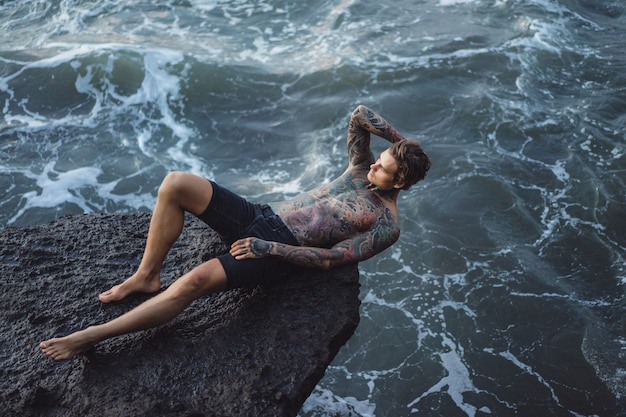 A tattooed man lies on the edge of a cliff. splashes of ocean waves.