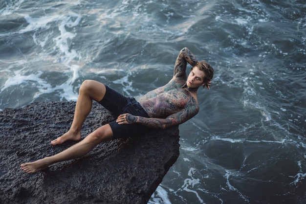 Un uomo tatuato giace sul bordo di una scogliera. schizzi di onde dell'oceano.