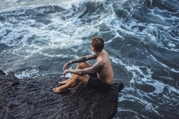 A tattooed man lies on the edge of a cliff. splashes of ocean waves.