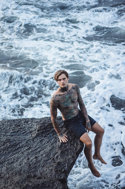 A tattooed man lies on the edge of a cliff. splashes of ocean waves.