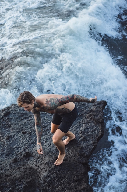 A tattooed man lies on the edge of a cliff. splashes of ocean waves.