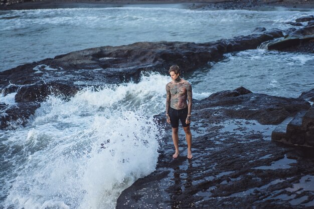 A tattooed man lies on the edge of a cliff. splashes of ocean waves.