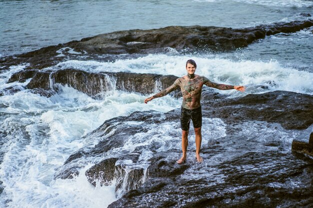 A tattooed man lies on the edge of a cliff. splashes of ocean waves.