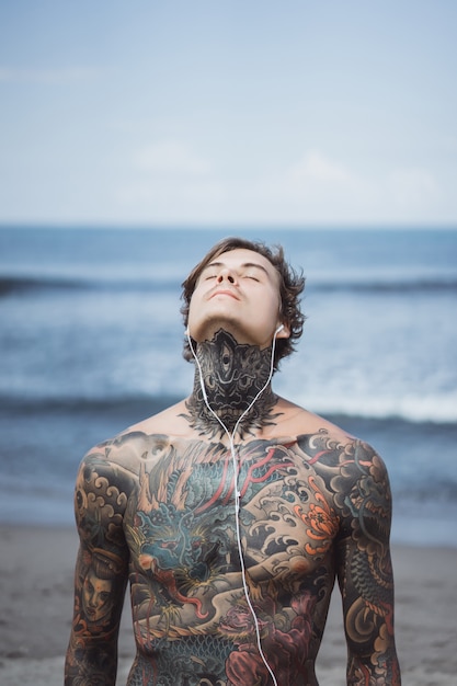 Foto gratuita uomo tatuato con le cuffie contro il cielo blu sull'oceano