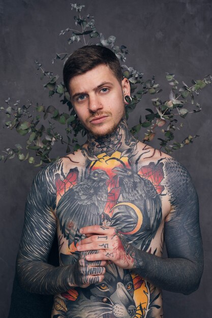 Татуированный мужчина с пирсингом в ушах и носу стоит за сухим растением на сером фоне