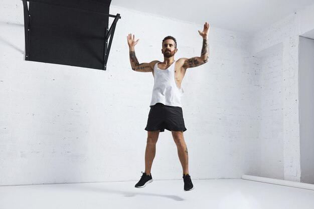 Tattooed and muscular athlete doing jumping jacks isolated on white brick wall next to black pull bar, looking left side