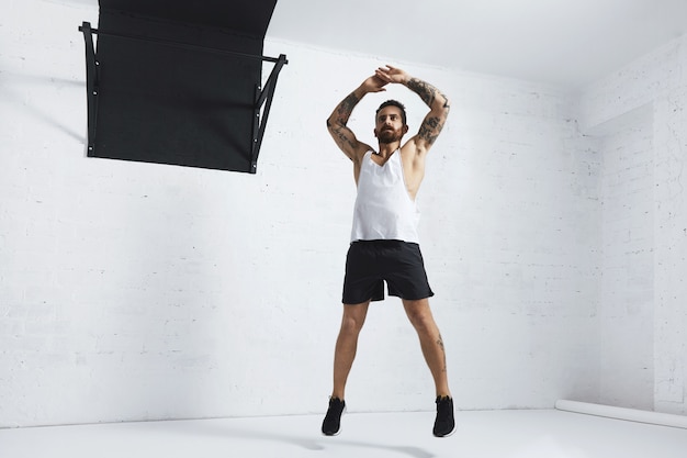 Foto gratuita atleta tatuato e muscoloso facendo jumping jacks isolato sul muro di mattoni bianco accanto alla barra di trazione nera