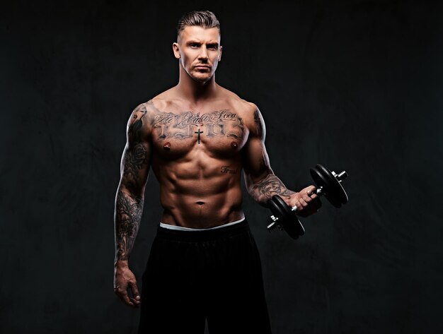 A tattooed muscular man doing exercises with dumbbells for biceps on dark background.