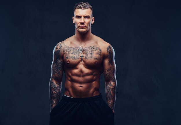 A tattooed muscular shirtless man with stylish hair posing at the camera over dark background.