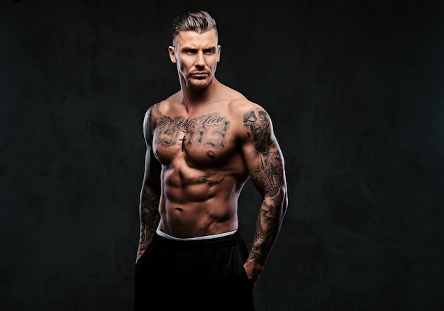 A tattooed muscular shirtless man with stylish hair posing at the camera on a dark background.