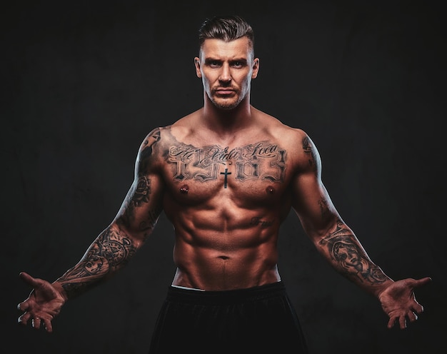 A tattooed muscular shirtless man with stylish hair posing at the camera on a dark background.