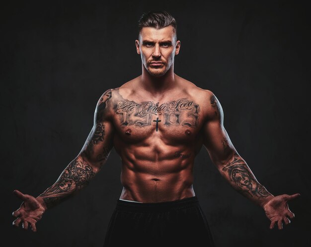 A tattooed muscular shirtless man with stylish hair posing at the camera on a dark background.