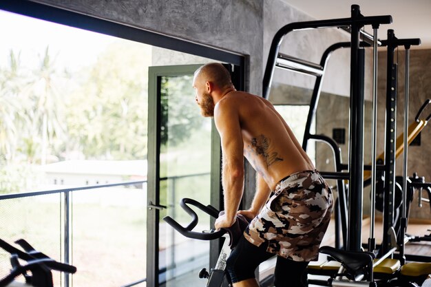 Tattooed muscular strong bearded man work out cardio on bicycle in gym near big window with view on trees outside