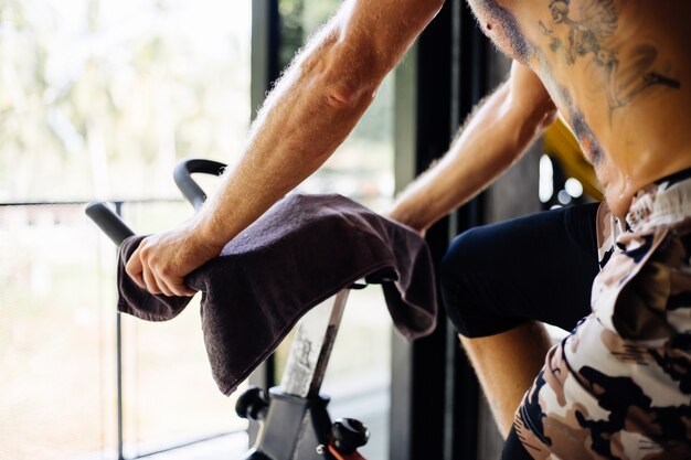 Татуированный мускулистый сильный бородатый мужчина тренируется кардио на велосипеде в тренажерном зале возле большого окна с видом на деревья снаружи