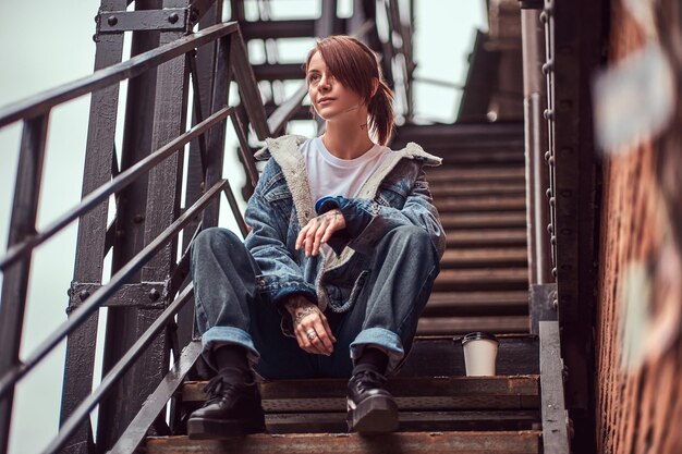 A tattooed redhead girl wearing trendy clothes sitting on stairs outside.