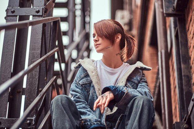 A tattooed redhead girl wearing trendy clothes sitting on stairs outside.