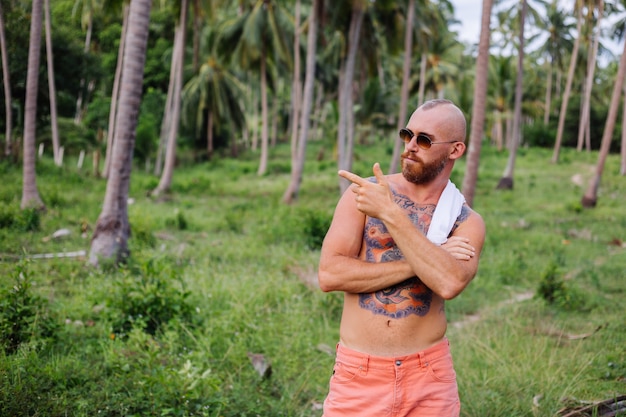 Tattooed strong man on jungle tropical field without shirt