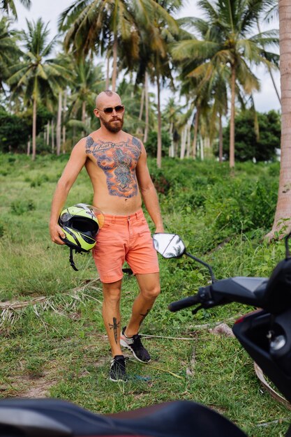 Tattooed strong man on tropical jungle field with red motorbike