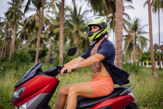 Татуированный сильный мужчина на поле тропических джунглей с красным мотоциклом