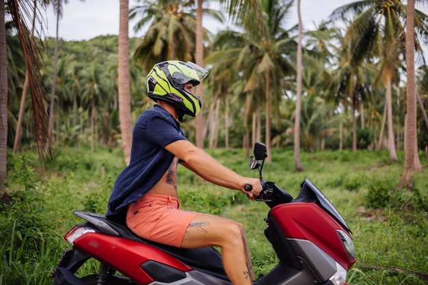 Татуированный сильный мужчина на поле тропических джунглей с красным мотоциклом