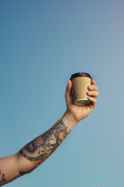 入れ墨の白人男性は、澄んだ青い空を背景にベージュの使い捨てコーヒーカップを高く保持します