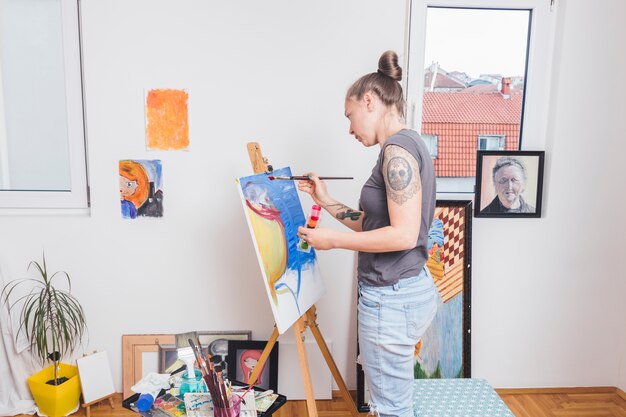 Tattooed woman painting colorful picture standing by window 