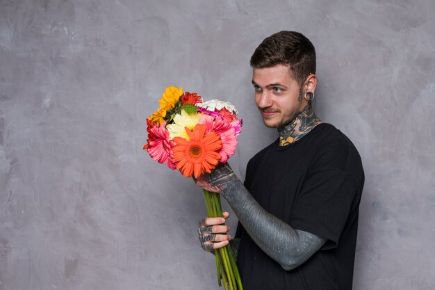 Tattooed young man looking at colorful gerbera flower bouquet standing against grey wall