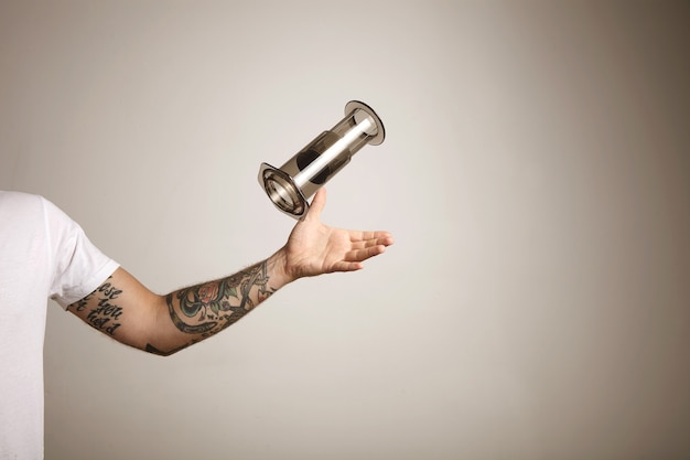 A tattooed young man in white t-shirt catching an empty gray aeropress on light gray wall