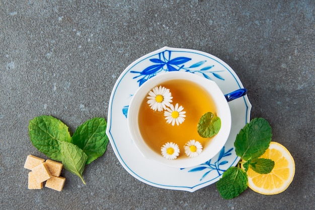 カップとソーサーにカモミールのお茶、レモン、ブラウンシュガーキューブ、緑の葉の灰色の漆喰の背景に平面図