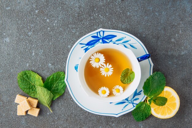 Tea of chamomile in a cup and saucer with lemon, brown sugar cubes and green leaves top view on a grey stucco background