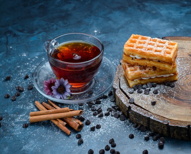 Tea in a cup with cinnamon sticks, waffle, choco chips, flowers, wooden board high angle view on a blue surface