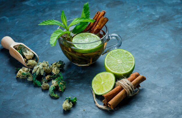 Tea in cup with mint, cinnamon, dried herbs, lime on grungy blue surface, high angle view.