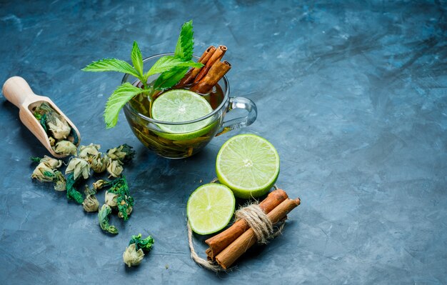 Tea in cup with mint, cinnamon, dried herbs, lime on grungy blue surface, high angle view.