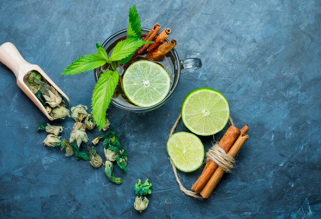 Tea in cup with mint, cinnamon, dried herbs, lime on grungy blue surface, top view.