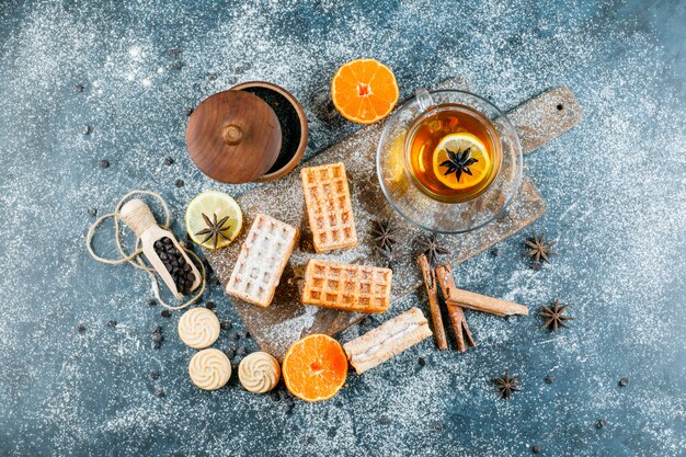 Tea in cup with waffle, biscuit, spices, choco chips, orange top view on grungy and cutting board