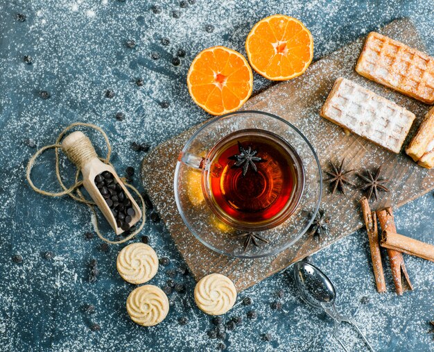 Tea in a cup with waffle, biscuit, spices, choco chips, strainer, orange flat lay on blue and cutting board