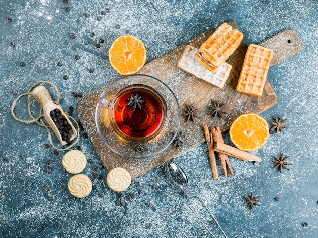 Tea in a cup with waffle, biscuit, spices, choco chips, strainer, orange top view on blue and cutting board