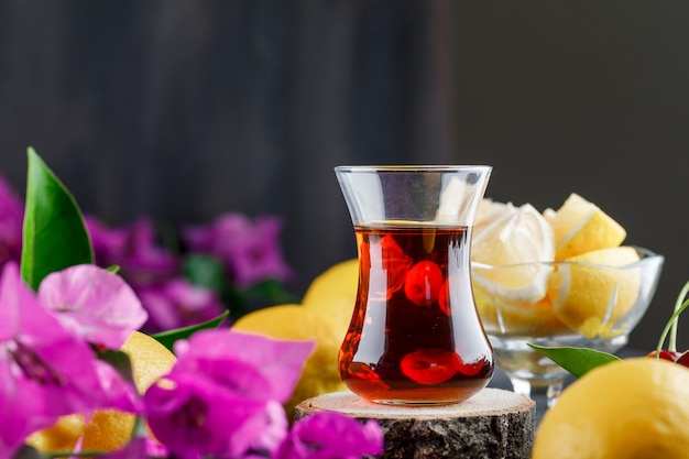 Tea in glass with lemons and slices, flowers, wooden board side view on a dark surface