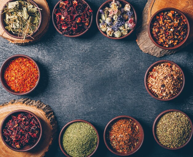 Tea herbs in a bowls with wood stubs flat lay on a dark textured background. space for text