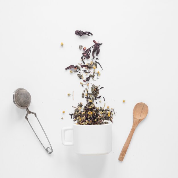 Tea herbs falling from cup with strainer and spoon on white backdrop