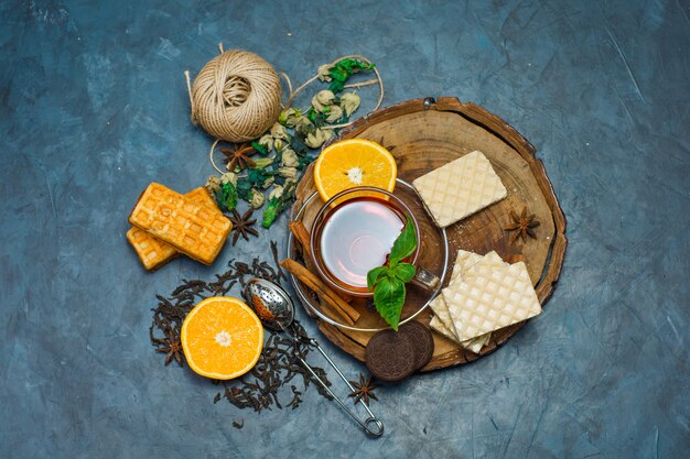 Tea in a mug with herbs, orange, spices, biscuits, thread, strainer top view on wooden board and stucco background