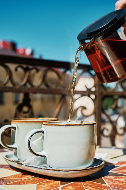 古いベランダでの朝のお茶の時間ガラスのティーポットの赤茶がカップに注がれます垂直ショット夏休みのお茶会の雰囲気休息とリラクゼーションカップに選択的な焦点