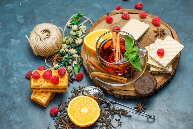 Tea with herbs, fruits, spices, biscuits, strainer, thread in a mug on wooden board and stucco background, flat lay.