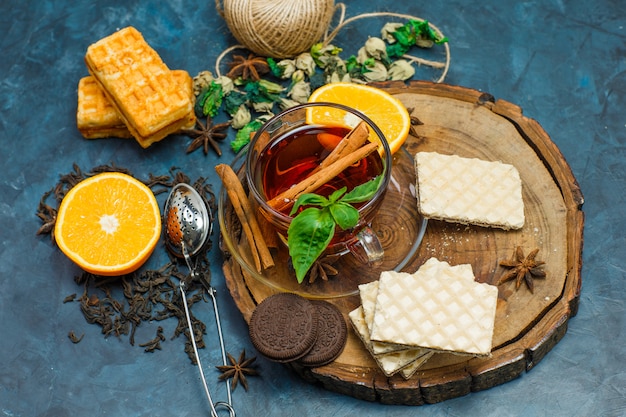 Tea with herbs, orange, spices, biscuits, strainer in a mug on wooden board and stucco background, flat lay.